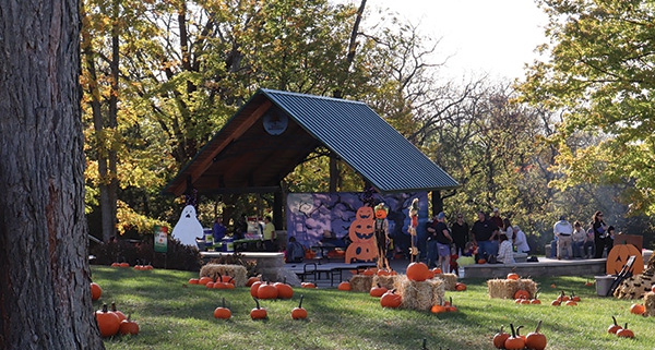 A group of people gather on a sunny autumn day at a park shelter decorated for Halloween, featuring jack-o-lanterns, ghost cutouts, and a scarecrow against a purple backdrop with a gnarled tree. The grounds are adorned with real pumpkins and hay bales.